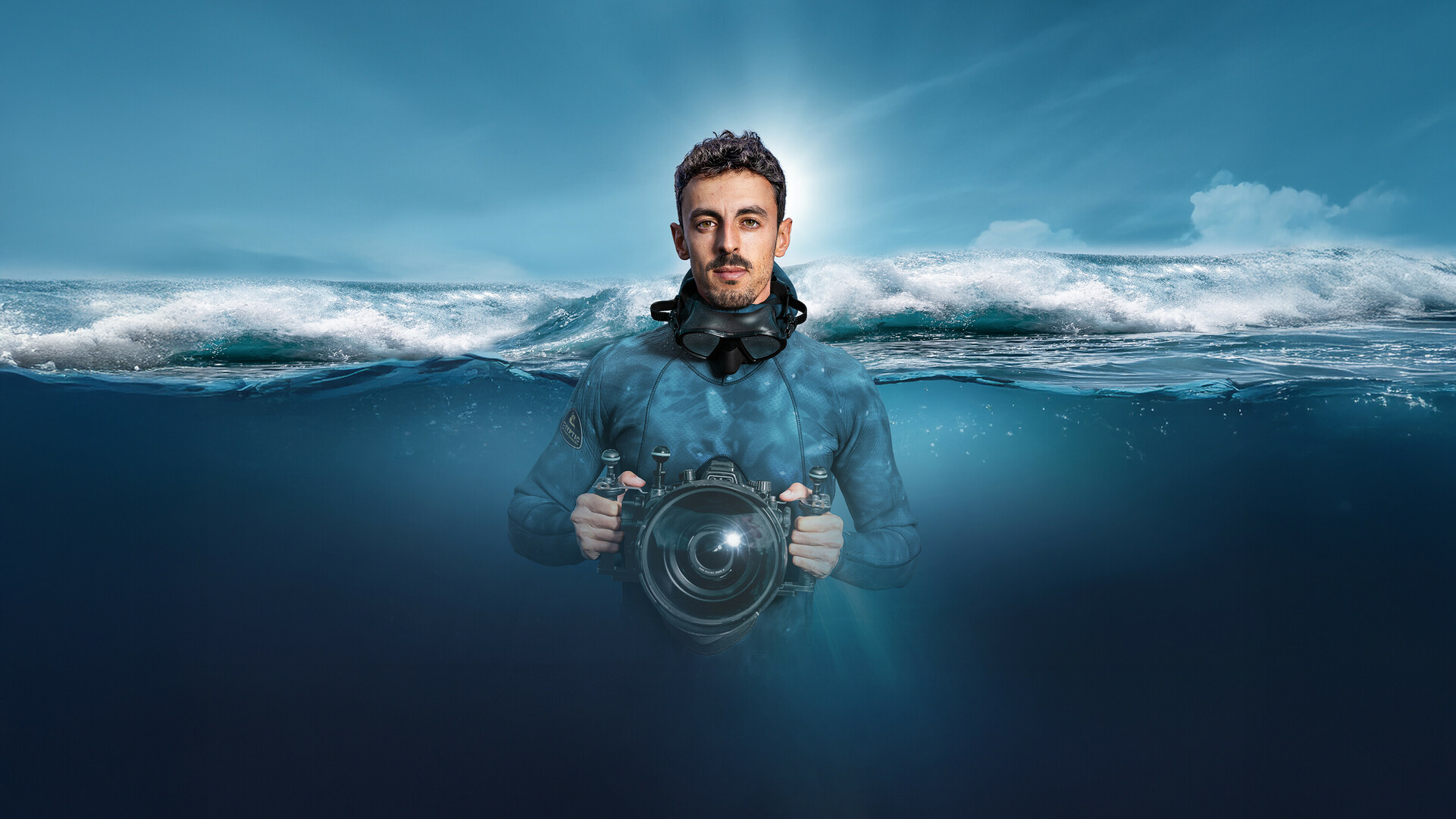 Diver with a diving mask and underwater camera in the ocean, half-submerged, with a breaking wave in the background.